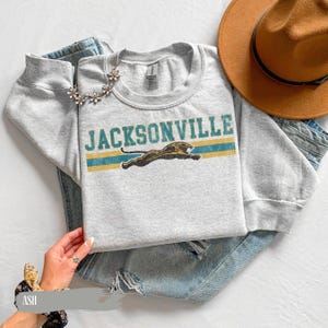 May include: A light gray crewneck sweatshirt featuring the text JACKSONVILLE in teal letters above a graphic of a leaping jaguar. The design includes two horizontal teal stripes. The sweatshirt is paired with blue denim jeans and a brown hat.
