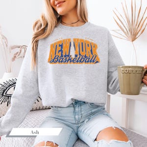 May include: A light gray sweatshirt with the text "NEW YORK Basketball" in orange and blue. The sweatshirt is a crew neck style and is made of a soft material. The word "Ash" is visible at the bottom of the image.