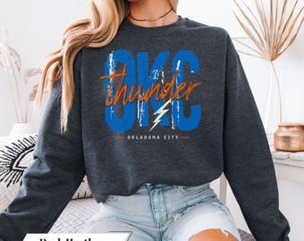 May include: Dark heather gray sweatshirt with the text "OKC Thunder" in blue and orange, with a lightning bolt graphic. The words "Oklahoma City" are below the graphic. The sweatshirt is a dark gray color.