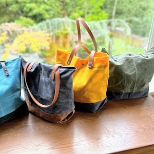 Peut inclure: Quatre sacs fourre-tout en toile cirée avec des poignées en cuir. Les sacs sont de couleur turquoise, gris foncé, jaune et vert olive. Chaque sac présente une base foncée contrastante, des rivets métalliques et des anneaux en D pour les anses.