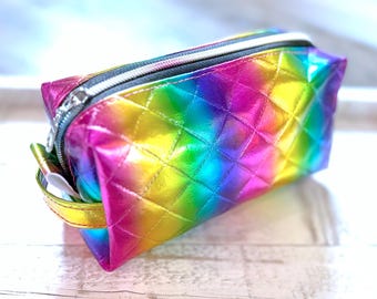 May include: A rainbow-colored, quilted cosmetic bag with a metallic sheen. The bag features a silver zipper closure and a small, gold-colored handle. The bag's vibrant colors transition from pink to yellow, green, and blue.