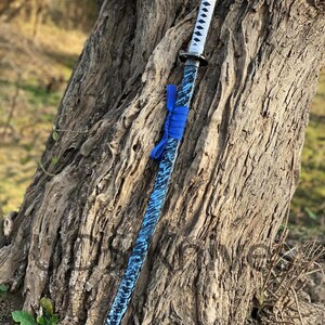 May include: A katana sword with a blue and black patterned blade and a white handle. The sword is adorned with a blue ribbon near the hilt. The blade has a textured appearance, and the handle is wrapped in white material.