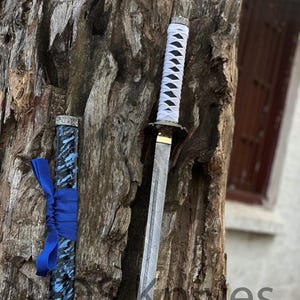 May include: A katana sword with a white wrapped handle and a patterned blade. The scabbard is black and blue with a blue ribbon tied around it. The sword is displayed against a textured tree trunk.