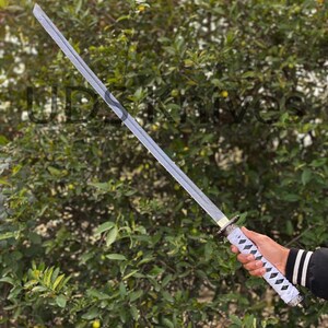 May include: A silver-bladed katana sword with a white and black diamond-patterned handle. The sword is held against a backdrop of green foliage. The text "UDSknives" is partially visible.
