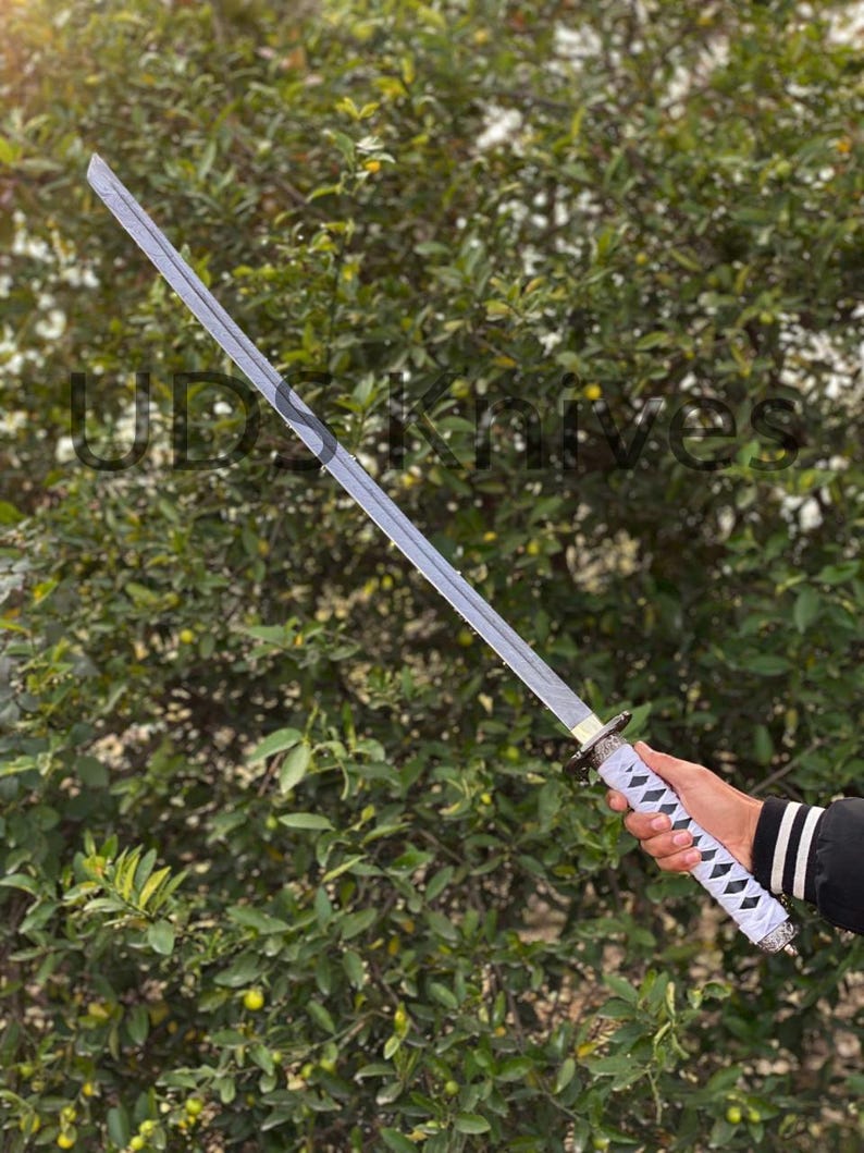 May include: A silver-bladed katana sword with a white and black diamond-patterned handle. The sword is held against a backdrop of green foliage. The text "UDSknives" is partially visible.