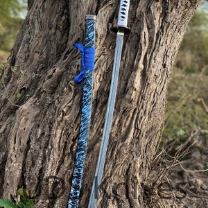 May include: A katana sword with a silver blade and white handle, next to its scabbard. The scabbard is blue with a black and white pattern and a blue ribbon. The sword is leaning against a tree trunk.