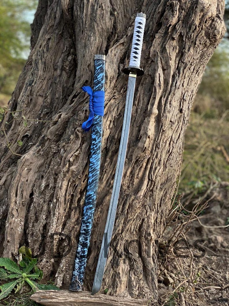May include: A katana sword with a silver blade and white handle, next to its scabbard. The scabbard is blue with a black and white pattern and a blue ribbon. The sword is leaning against a tree trunk.