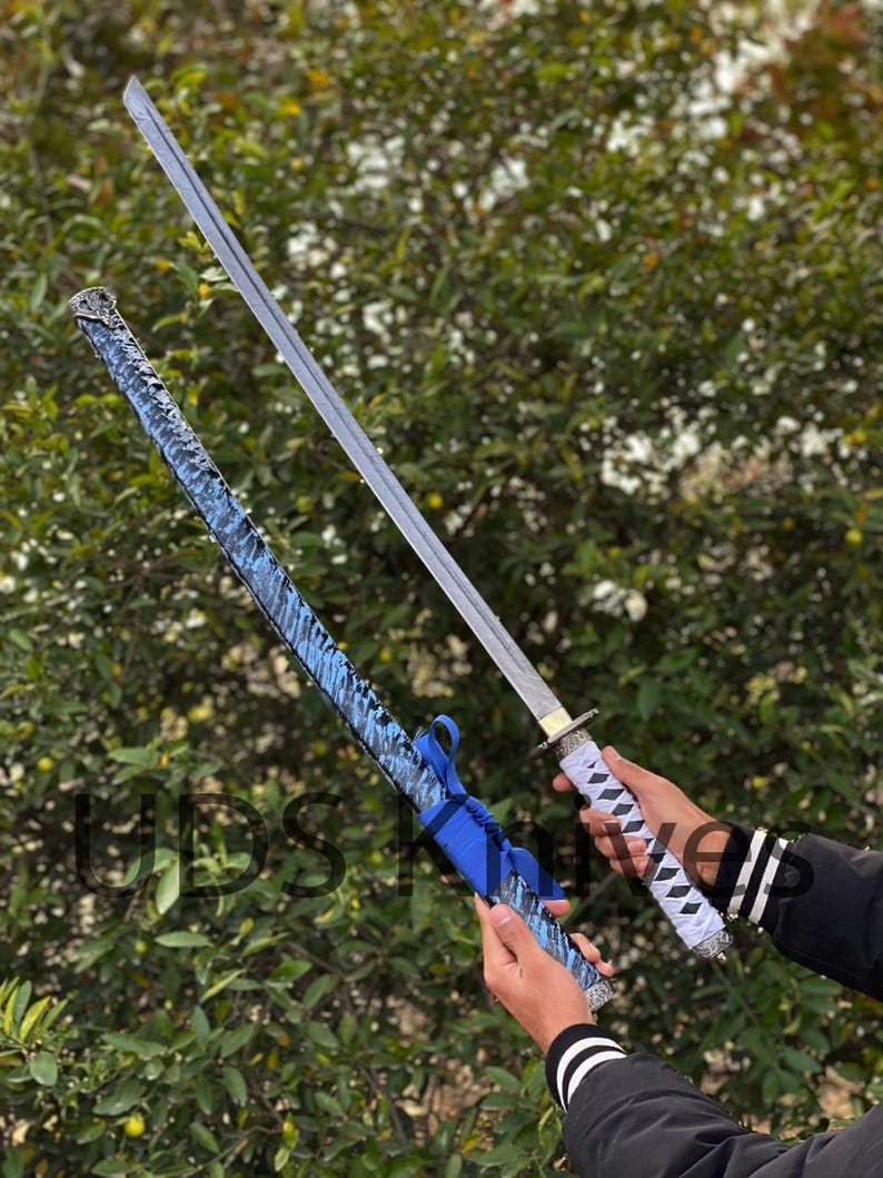 May include: A katana sword with a silver blade and white and black handle is held next to its blue patterned scabbard. The scabbard has a blue ribbon tied around it. The background is a green bush.
