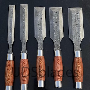 Könnte beinhalten: Satz von fünf Holzmeißeln mit gemusterten Stahlklingen und Holzgriffen. Die Griffe sind warmbraun mit einer silbernen Zwinge. Die Klingen haben ein einzigartiges strukturiertes Muster. Die Meißel sind vertikal angeordnet.