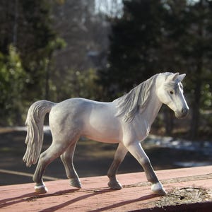 May include: A detailed figurine of a white horse with a gray mane and tail. The horse is posed in a walking stance, with its legs in motion. The figurine is set against a blurred background of trees.