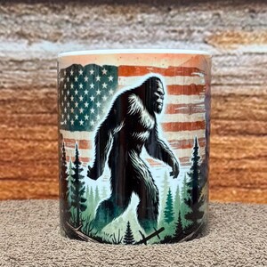 May include: A cylindrical coffee mug featuring a Bigfoot illustration against a backdrop of the American flag and a forest scene. The mug is predominantly white with a wraparound design, showcasing the creature in black and the flag in red, white, and blue.