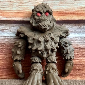 May include: A brown Bigfoot figurine with red eyes. The figure has a detailed, furry texture and articulated joints. The creature stands upright with large feet and hands, and a stern expression. The background is a textured, wood-like surface.