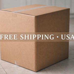May include: A large, unopened cardboard box, sealed with tape. The box is a light brown color and sits on a tiled floor. The text "FREE SHIPPING &bull; USA" is overlaid on the box.