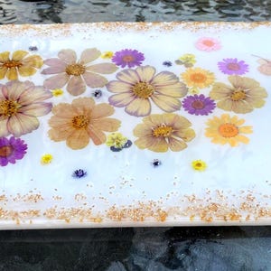 May include: A rectangular white resin tray with gold handles and gold glitter trim. The tray is decorated with dried flowers in shades of yellow, purple, and pink. The flowers are embedded in the resin, creating a decorative serving piece.