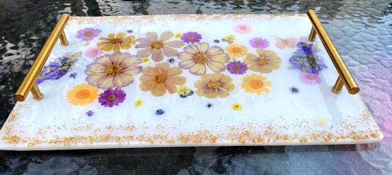May include: A rectangular white resin tray with gold handles and gold glitter trim. The tray is decorated with dried flowers in shades of yellow, purple, and pink. The flowers are embedded in the resin, creating a decorative serving piece.