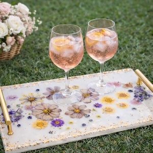 May include: A decorative serving tray with dried flowers embedded in a white resin surface. Two wine glasses filled with a pink beverage and ice sit on the tray. Gold handles and a basket of flowers are in the background.