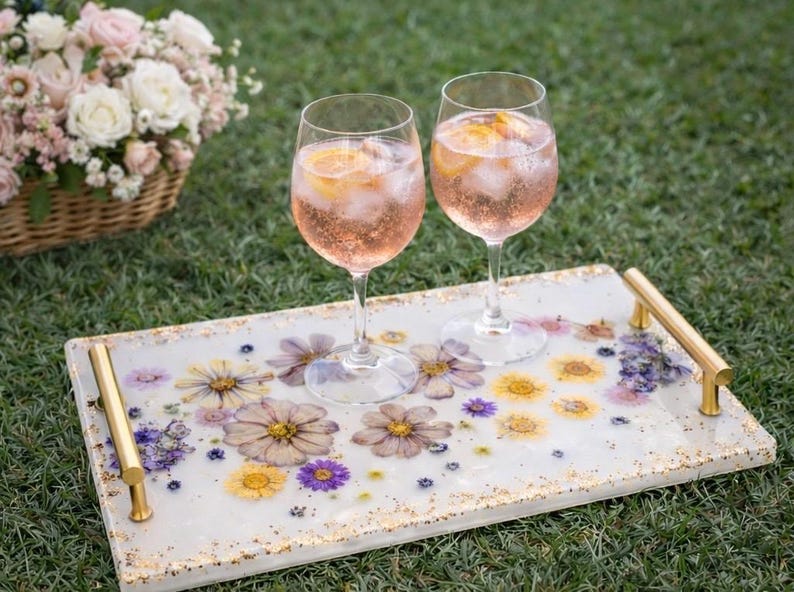 May include: A decorative serving tray with dried flowers embedded in a white resin surface. Two wine glasses filled with a pink beverage and ice sit on the tray. Gold handles and a basket of flowers are in the background.