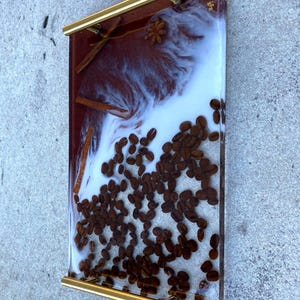 May include: A rectangular resin tray with gold handles, featuring a coffee theme. The tray incorporates coffee beans, cinnamon sticks, and star anise within a clear resin, with a white and brown marbled effect. The tray measures approximately 12 inches long and 8 inches wide.