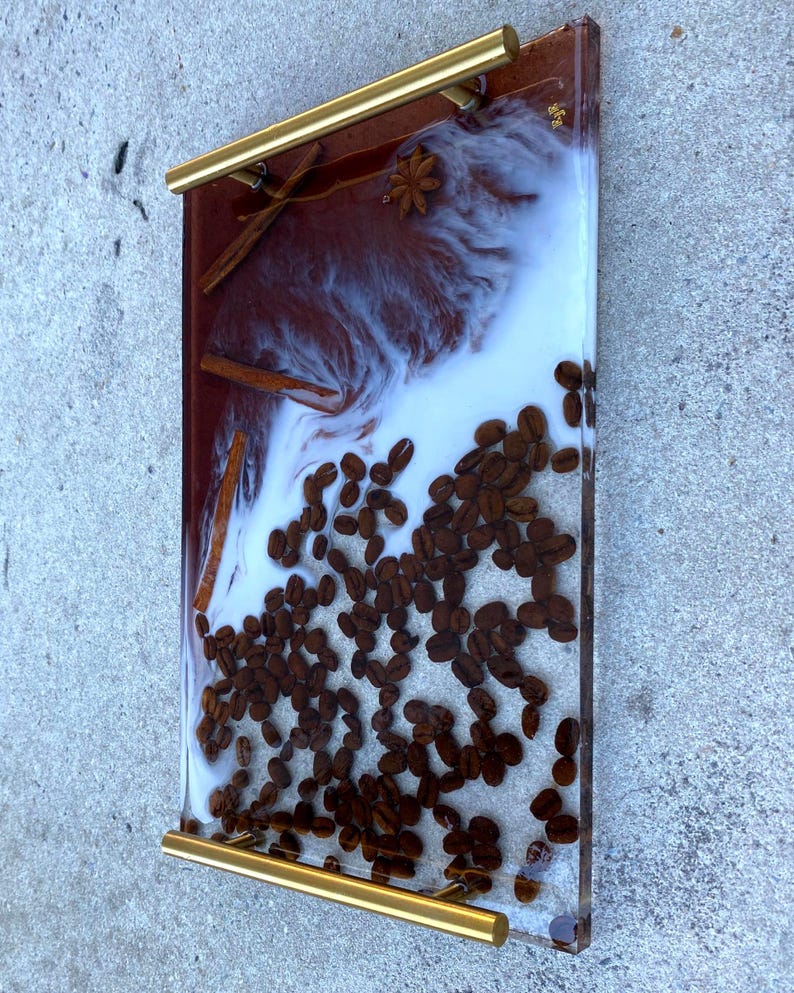 May include: A rectangular resin tray with gold handles, featuring a coffee theme. The tray incorporates coffee beans, cinnamon sticks, and star anise within a clear resin, with a white and brown marbled effect. The tray measures approximately 12 inches long and 8 inches wide.