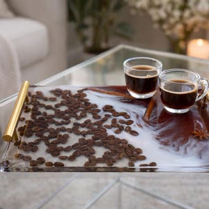 May include: A rectangular serving tray with gold handles, filled with coffee beans and a white and brown resin design. Two clear glass cups of coffee sit on the tray. A decorative and functional item for serving.