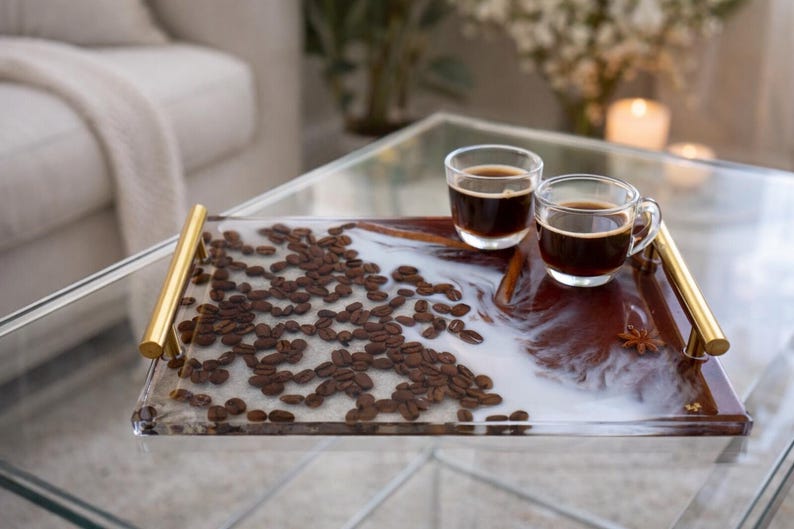 May include: A rectangular serving tray with gold handles, filled with coffee beans and a white and brown resin design. Two clear glass cups of coffee sit on the tray. A decorative and functional item for serving.