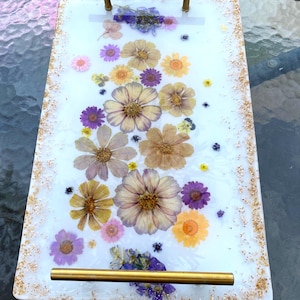 May include: A rectangular serving tray with a white surface embedded with dried flowers in shades of yellow, purple, and pink. The tray has gold-colored handles and gold flecks along the edges. A decorative piece for serving or display.