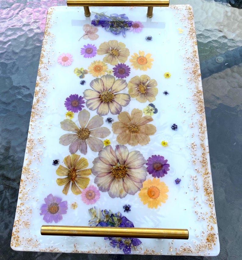 May include: A rectangular serving tray with a white surface embedded with dried flowers in shades of yellow, purple, and pink. The tray has gold-colored handles and gold flecks along the edges. A decorative piece for serving or display.