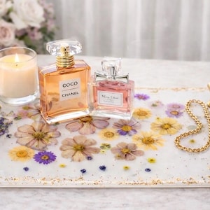May include: A decorative tray with gold handles, featuring embedded dried flowers and gold flecks. Two perfume bottles and a lit candle sit on the tray. One perfume bottle is labeled "COCO CHANEL".