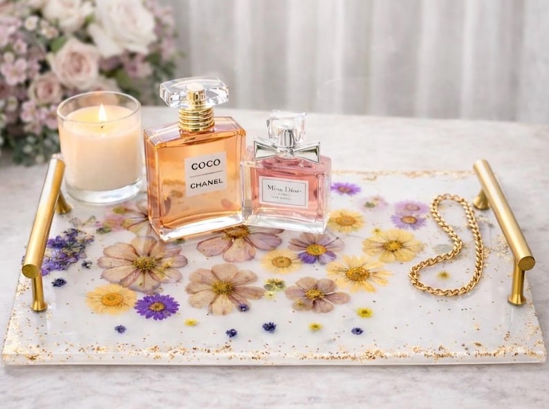 May include: A decorative tray with gold handles, featuring embedded dried flowers and gold flecks. Two perfume bottles and a lit candle sit on the tray. One perfume bottle is labeled "COCO CHANEL".