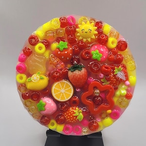 May include: A circular resin coaster featuring a translucent base embedded with various colorful plastic charms including strawberries, lemon slices, a yellow sun, a red star, and numerous round beads in shades of red, pink, yellow, and orange.