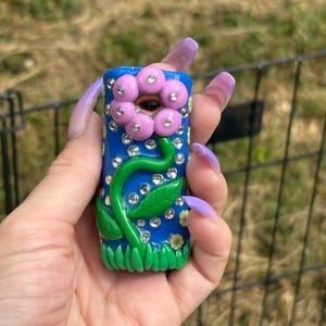 May include: A blue, rectangular, decorative lighter featuring a pink flower design with rhinestones. The flower has pink petals and a black center. A green stem and leaves are also present. The lighter is held in a hand with long, purple nails.