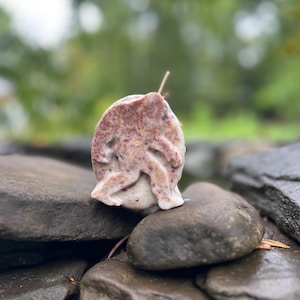 May include: A handcrafted soap shaped like the mythical Bigfoot, with a textured, speckled appearance. The soap is primarily a light pink color with darker pink and white accents, resting on a bed of dark gray stones.