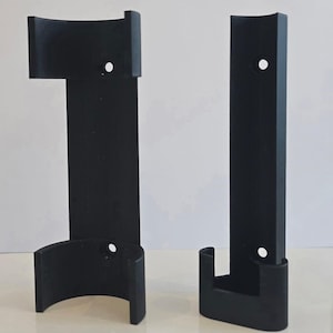 Könnte beinhalten: Zwei schwarze Wandhalterungen. Eine Halterung hat eine gebogene Ober- und Unterseite mit einer vertikalen Stütze. Die andere Halterung ist eine einfache, vertikale Stütze mit gebogenem Boden. Beide haben ein Loch.