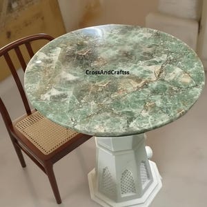 May include: A round table with a green marble top and a white geometric base. A wooden chair with a woven seat is next to the table. The text "CrossAndCraftss" is visible on the table's surface.
