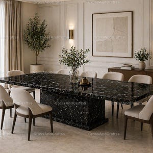 Op de afbeelding: Een moderne eetkamer met een grote zwarte marmeren eettafel met witte adering. De tafel wordt omringd door acht beige gestoffeerde stoelen. De kamer heeft beige muren, een groot ingelijst kunstwerk en decoratieve planten.
