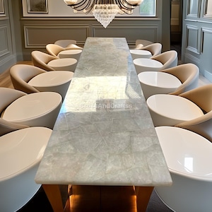 Puede incluir: Una mesa de comedor larga y rectangular con una superficie texturizada de color gris claro. La mesa está rodeada de sillas beige con respaldos redondeados y mesas auxiliares blancas y circulares. Una lámpara de araña de cristal cuelga sobre la mesa.