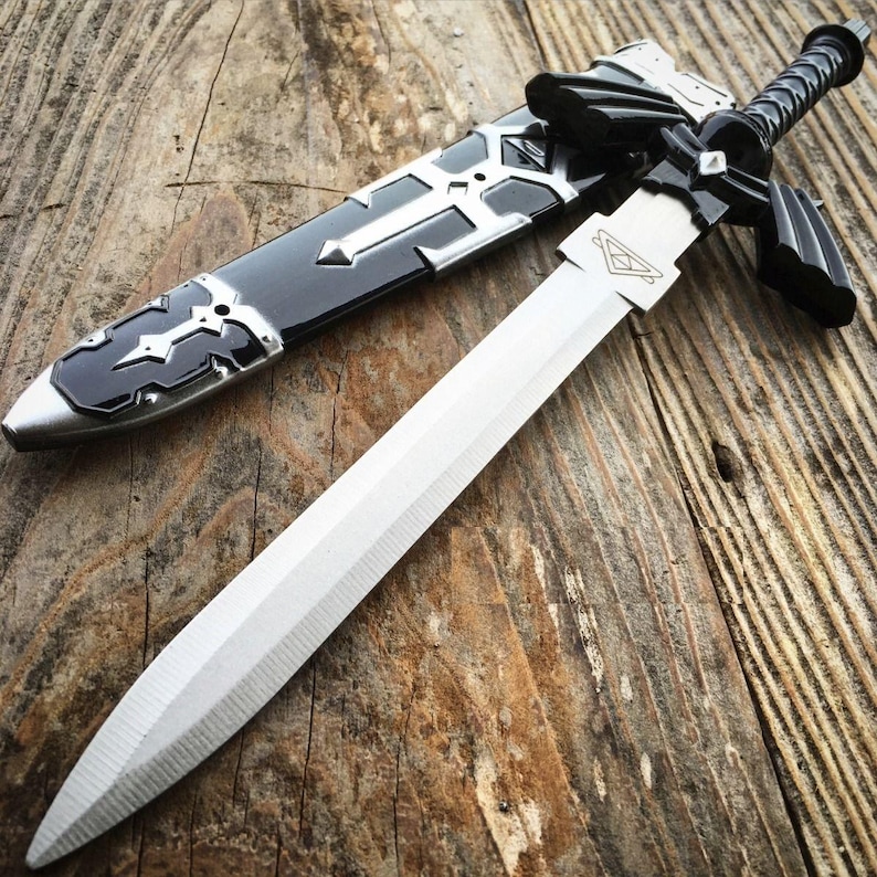 May include: A silver and black sword with a matching sheath. The sword has a long, straight blade and a black handle. The sheath is black with silver accents. The sword is lying on a wooden surface.