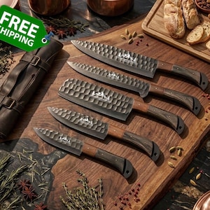 May include: A set of five kitchen knives with hammered metal blades and dark wood handles. Each blade features a mountain logo engraving. The knives are arranged in a row on a wooden surface, accompanied by a brown leather knife roll case.