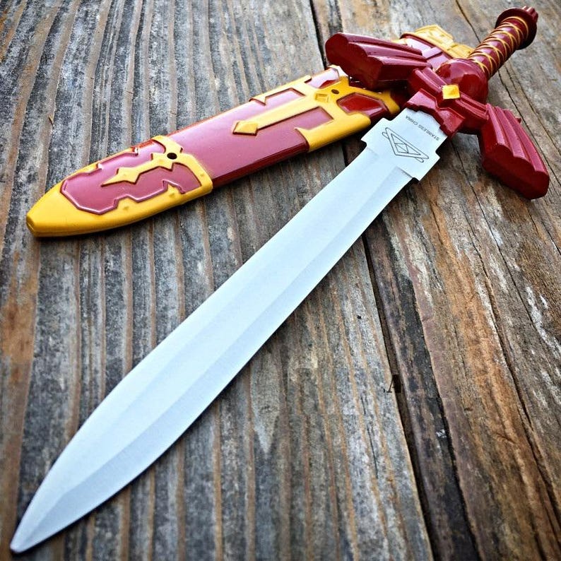 May include: A decorative sword with a silver blade and a red and gold hilt and scabbard. The hilt features a diamond-shaped design. The scabbard is red with gold accents. The sword is lying on a wooden surface.