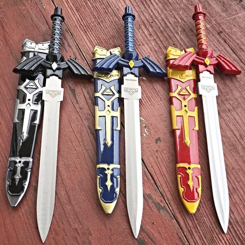 May include: Three toy swords with sheaths in black, blue, and red. Each sword has a silver blade and a decorative hilt. The sheaths are detailed with gold accents and geometric designs. The swords are displayed on a wooden surface.