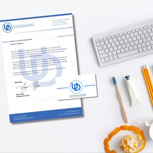 Peut inclure: Vue de dessus d'un espace de travail avec en-tête, carte de visite, clavier, crayons et brosse à dents. L'en-tête et la carte affichent le logo "Universaenix Design" et les coordonnées. Une orange à moitié mangée est à côté d'une tasse blanche.