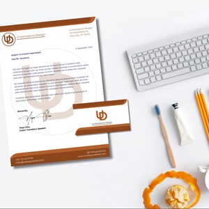Op de afbeelding: Een briefpapier set met briefhoofd en visitekaartje, beide met het "Universaenix Design" logo in bruin en wit. Een toetsenbord, potloden, een tandenborstel en een kopje staan ook op het witte oppervlak.