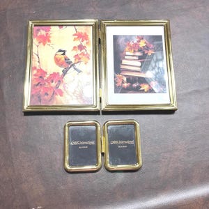 May include: A collection of gold-toned picture frames. Two larger frames display autumn-themed artwork: a bird on a branch and a stack of books with leaves. A smaller, hinged frame is also included, with the text "CARR International" visible.