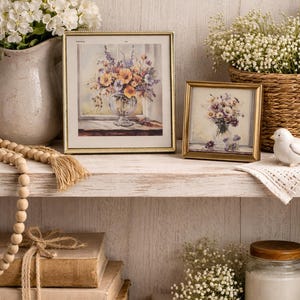 May include: A decorative shelf displays framed floral art, a vase of white flowers, and a woven basket with white flowers. Additional elements include a strand of wooden beads, stacked books, and a jar with a wooden lid.