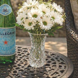 May include: A clear, cut-glass vase filled with white daisies and greenery sits on a dark metal table. A green bottle of sparkling water is to the left of the vase. The table has a decorative, openwork design.