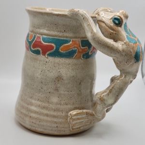 May include: A ceramic mug with a speckled off-white finish featuring a sculpted frog handle. The mug is decorated with a horizontal band of repeating teal and orange abstract shapes around the upper section, while the frog has teal accents.