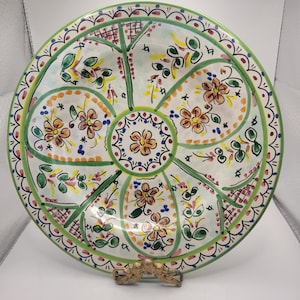 May include: A hand-painted ceramic serving platter with a floral design. The white plate features a green border and is divided into sections with orange, yellow, and blue floral patterns. The plate is supported by a gold-coloured metal stand.