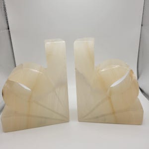 May include: A pair of off-white and cream-colored stone bookends featuring a smooth, polished finish with natural translucent veining. Each bookend has a vertical rectangular back and a curved, fan-like front section with carved geometric lines.
