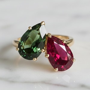 May include: A gold ring featuring two pear-cut gemstones set side by side. One gemstone is a deep forest green, while the other is a vibrant magenta red. The stones are held in place by small gold prongs on a thin, polished gold band.