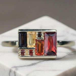 May include: A gold ring featuring a rectangular setting with a cluster of multi-coloured gemstones. The arrangement includes baguette-cut stones in deep red, yellow, orange, and dark brown, accented by small round red stones set in a metal frame.